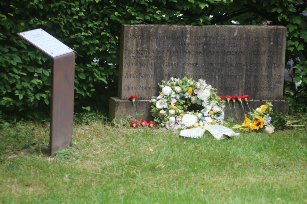 Gedenkstätte mit Blumenkranz und beschriftetem Schild.