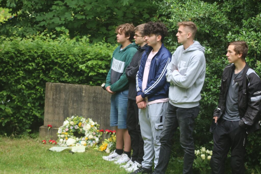 Gruppe von fünf Jugendlichen steht auf einem Friedhof neben Blumengebinden.