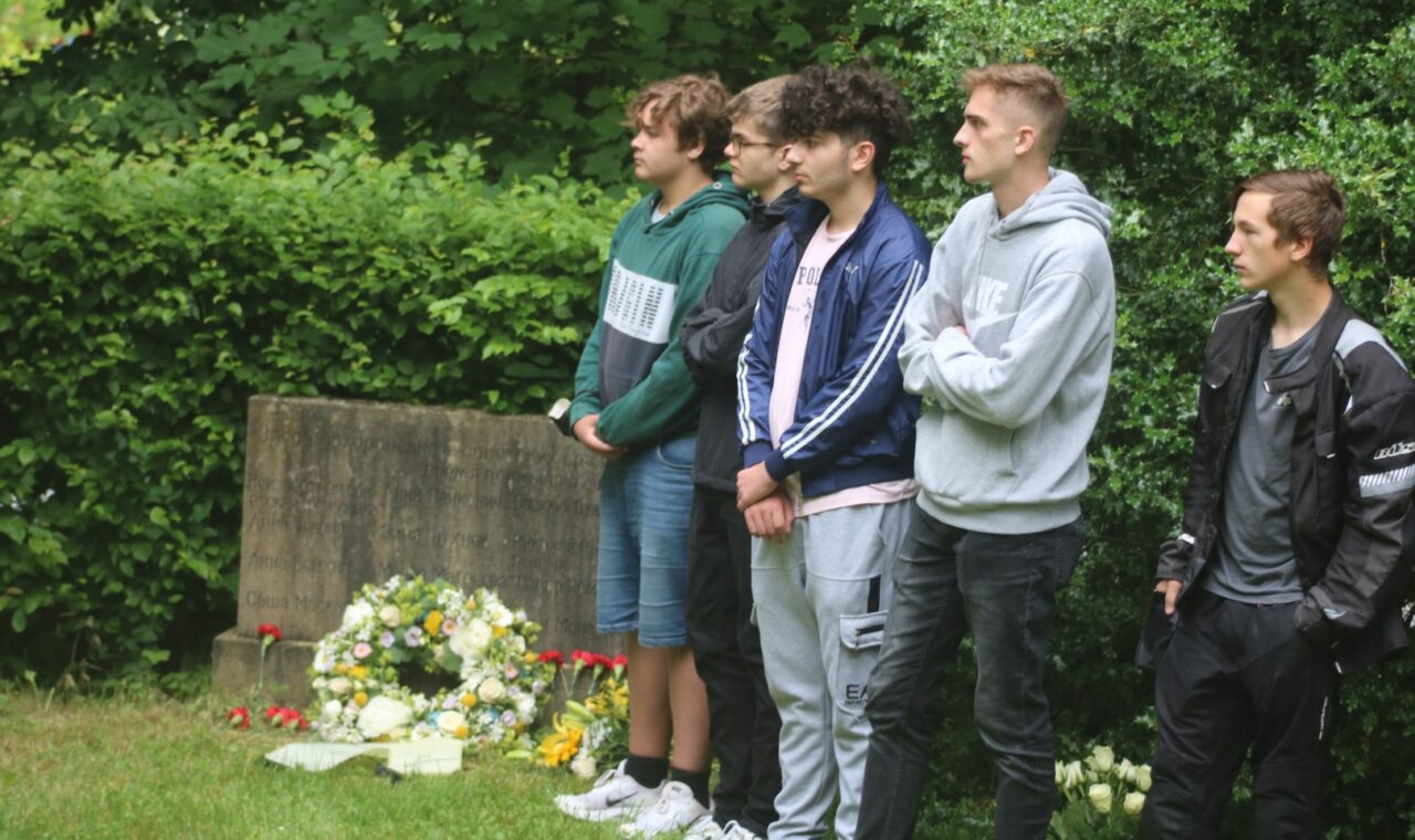 Gruppe von fünf Jugendlichen steht auf einem Friedhof neben Blumengebinden.