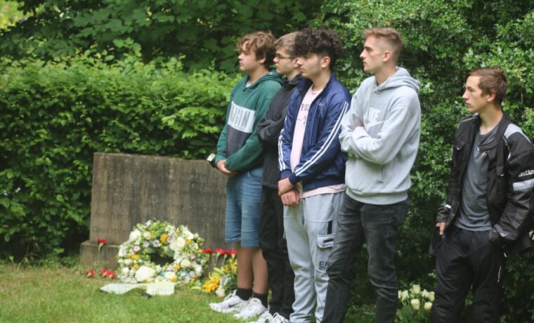 Gruppe von fünf Jugendlichen steht auf einem Friedhof neben Blumengebinden.