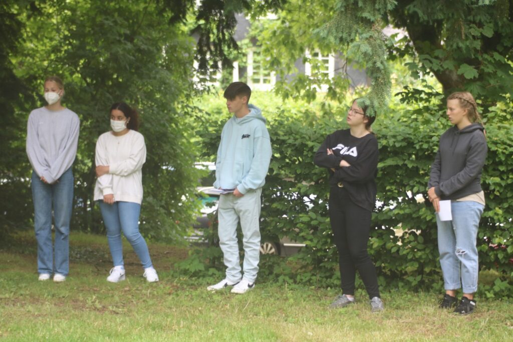 Sechs Jugendliche stehen im Freien und unterhalten sich. Einige tragen Masken.