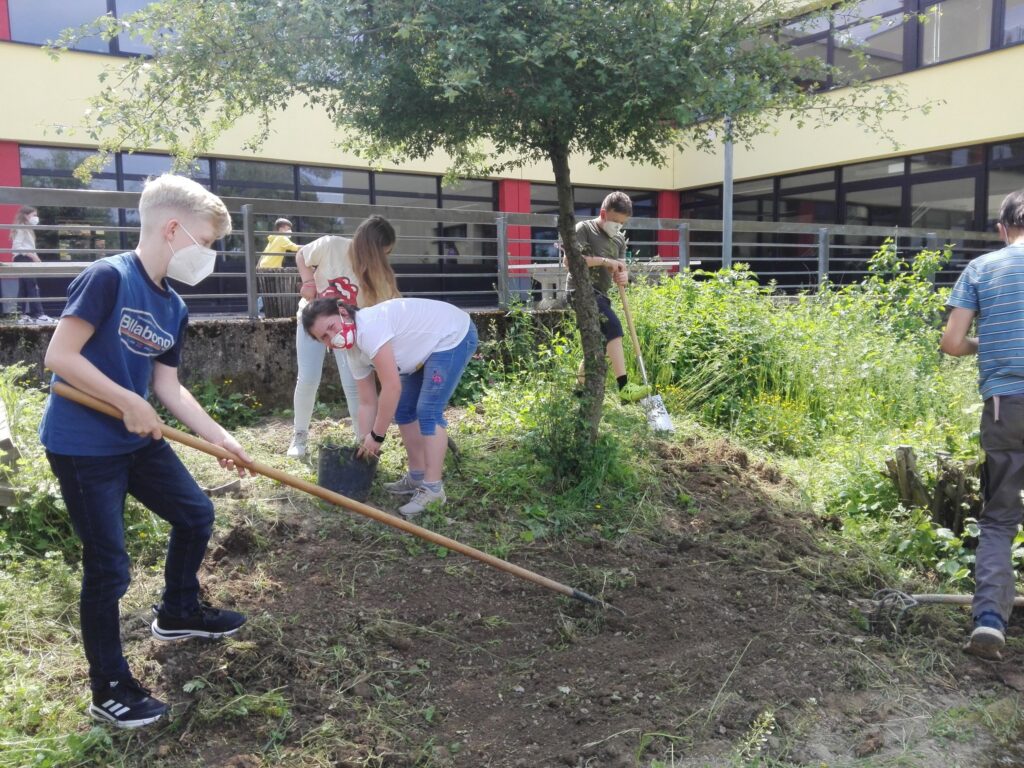 Schüler arbeiten im Garten und pflanzen unter einem Baum. Einige tragen Schutzmasken.