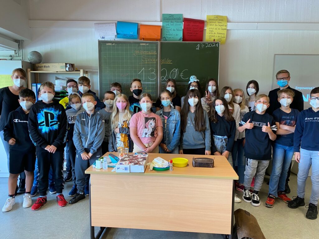 Schülergruppe steht in einem Klassenraum, alle tragen Masken. Tische mit Materialien sichtbar.