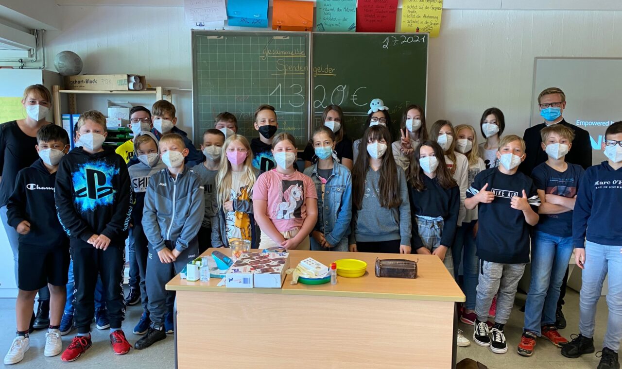Schülergruppe steht in einem Klassenraum, alle tragen Masken. Tische mit Materialien sichtbar.