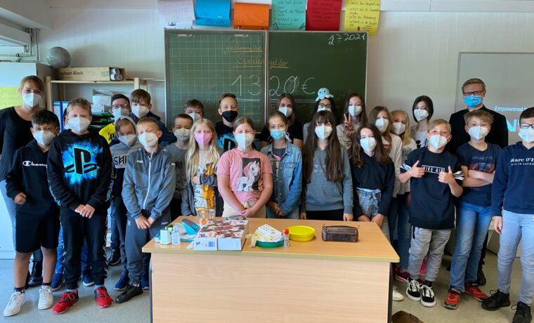 Schülergruppe steht in einem Klassenraum, alle tragen Masken. Tische mit Materialien sichtbar.