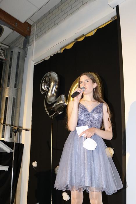 Mädchen in glitzerndem Kleid hält ein Mikrofon und spricht auf einer Bühne.