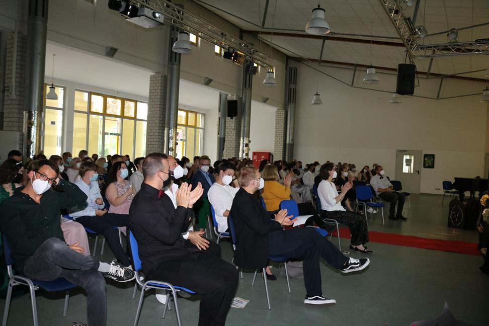 Menschen sitzen im Saal und applaudieren, einige tragen Mund-Nasen-Schutz.