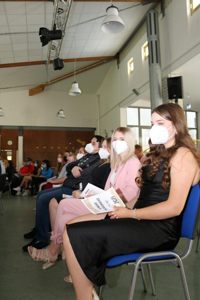 Schülerinnen und Schüler in Masken sitzen in einem Saal und hören zu.