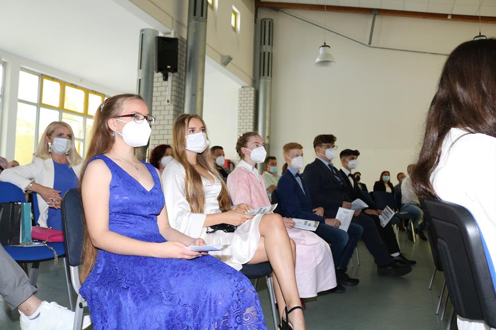 Schülerinnen und Schüler in festlicher Kleidung sitzen mit Masken im Veranstaltungsraum.