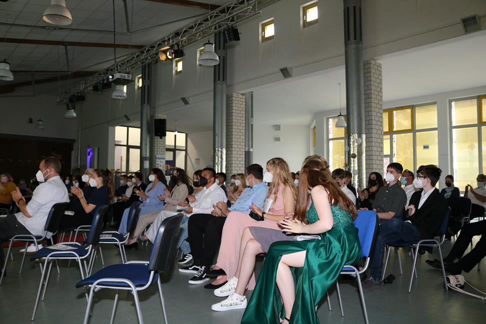 Menschen sitzen in einem Saal, viele tragen Masken. Blickrichtung nach vorne.