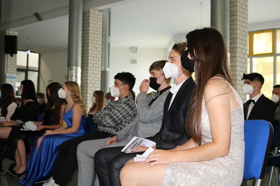Schüler in festlicher Kleidung sitzen in einem Raum und tragen Masken.