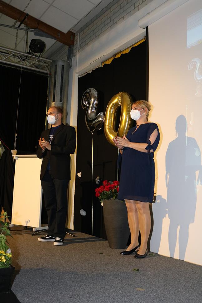 Zwei Personen auf einer Bühne mit Masken, dahinter Luftballons.