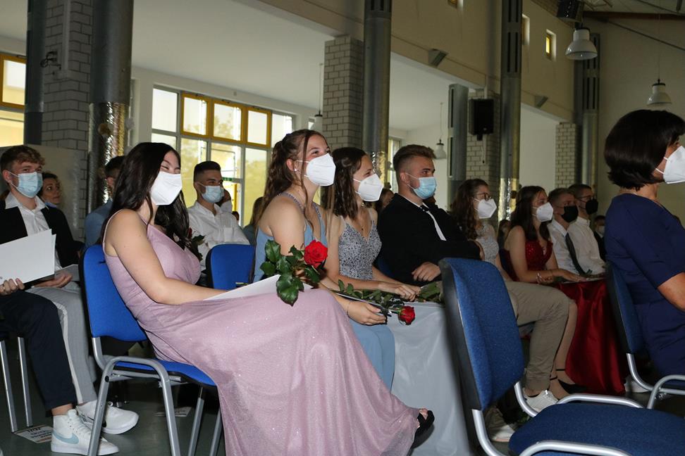 Schülerinnen und Schüler in festlicher Kleidung sitzen bei einer Feier und tragen Masken.