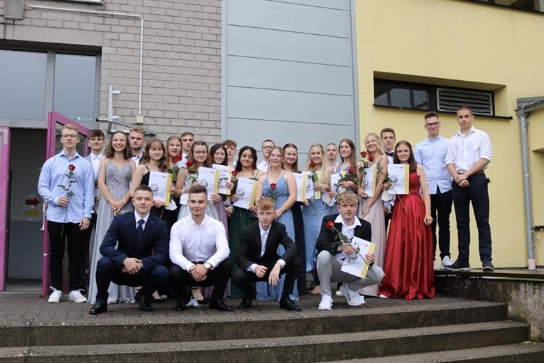 Gruppe von Schülern mit Urkunden und Blumen vor einem Schulgebäude.