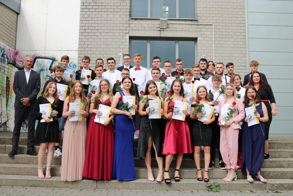 Gruppe von Schülern in festlicher Kleidung mit Zeugnissen und Blumen auf Treppen.