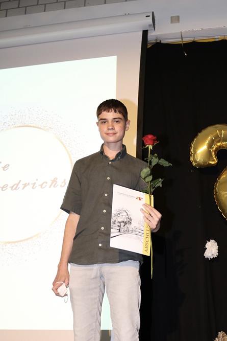 Ein junger Schüler hält eine Urkunde und eine Rose auf einer Bühne.