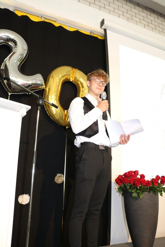 Ein junger Mann in Anzug hält ein Mikrofon vor einer Bühne mit Luftballons und Blumen.