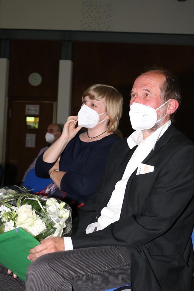 Zwei Menschen mit Mundschutz sitzen in einem Publikum, einer hält Blumen.