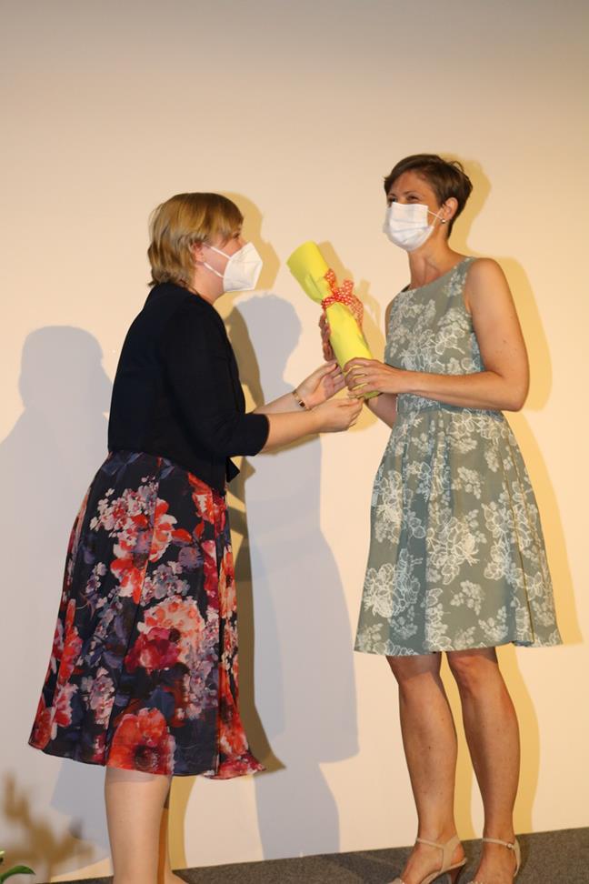Zwei Frauen überreichen sich eine Urkunde auf einer Bühne. Beide tragen Masken.