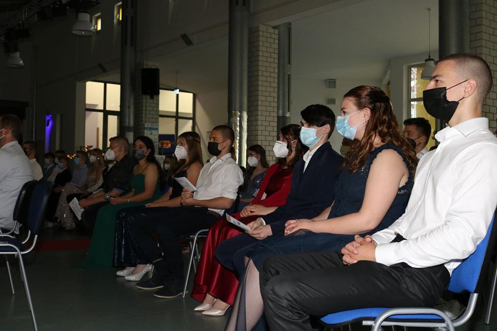 Gruppierung von Personen in Formeller Kleidung, die auf Stühlen sitzen und Masken tragen.