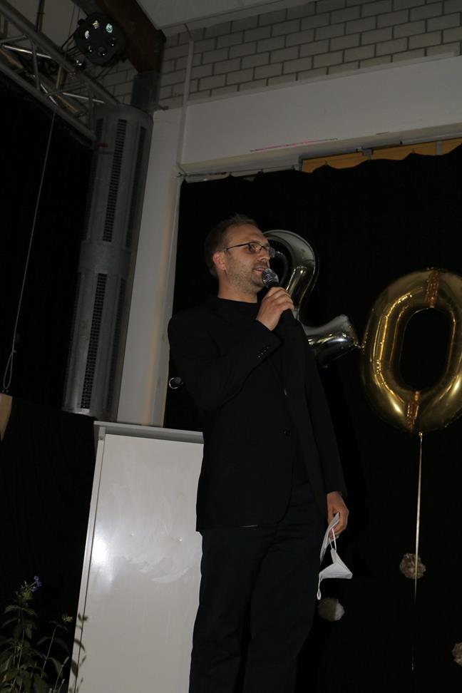 Mann spricht am Mikrofon, goldene Luftballons im Hintergrund.