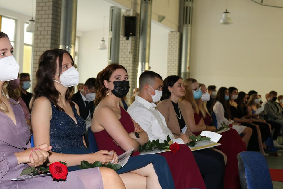 Absolventen sitzen in Reihen, tragen Masken und halten Blumen in der Hand.