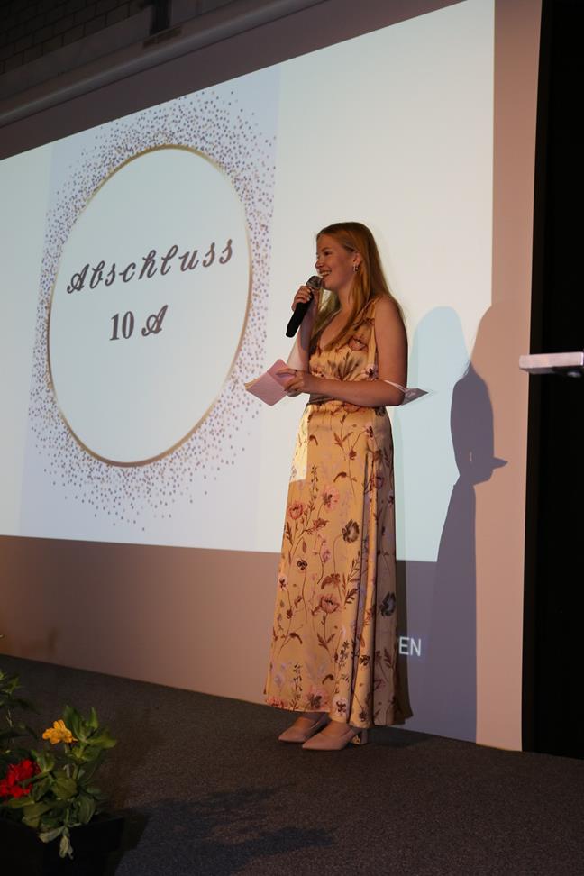 Frau mit Mikrofon steht vor einer Leinwand mit der Aufschrift „Abschluss 10a“.