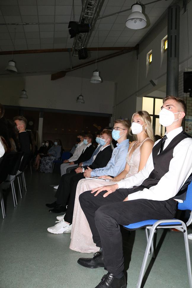 Schüler in festlicher Kleidung sitzen in einem Saal mit Masken.