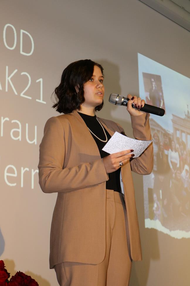 Person spricht mit Mikrofon auf einer Veranstaltung. Im Hintergrund sind Bilder zu sehen.