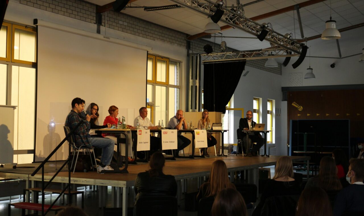 Podiumsdiskussion mit sechs Teilnehmern auf einer Bühne in einem Schulgebäude.