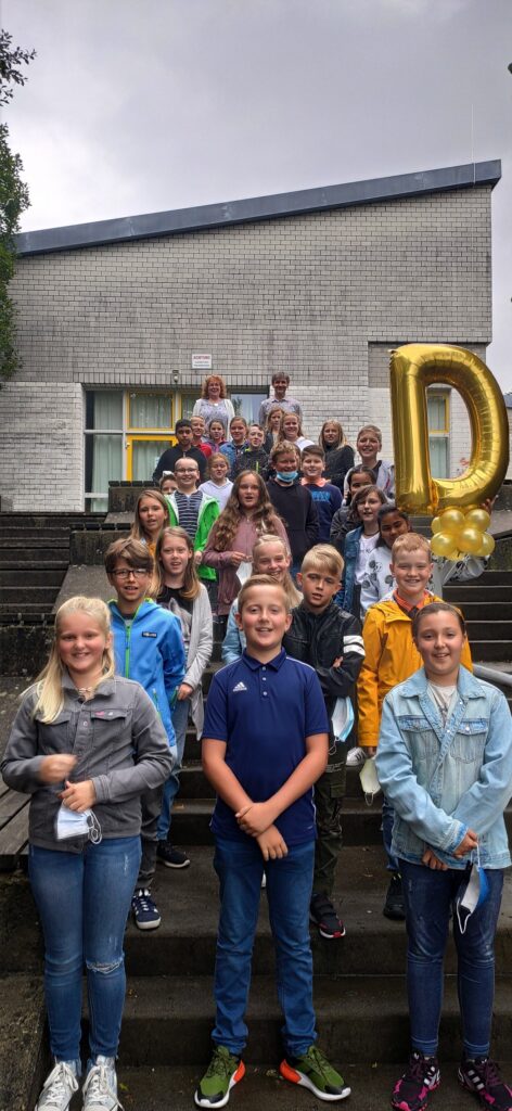 Gruppe von Schülern steht auf Stufen vor einem Schulgebäude, einige halten Luftballons.