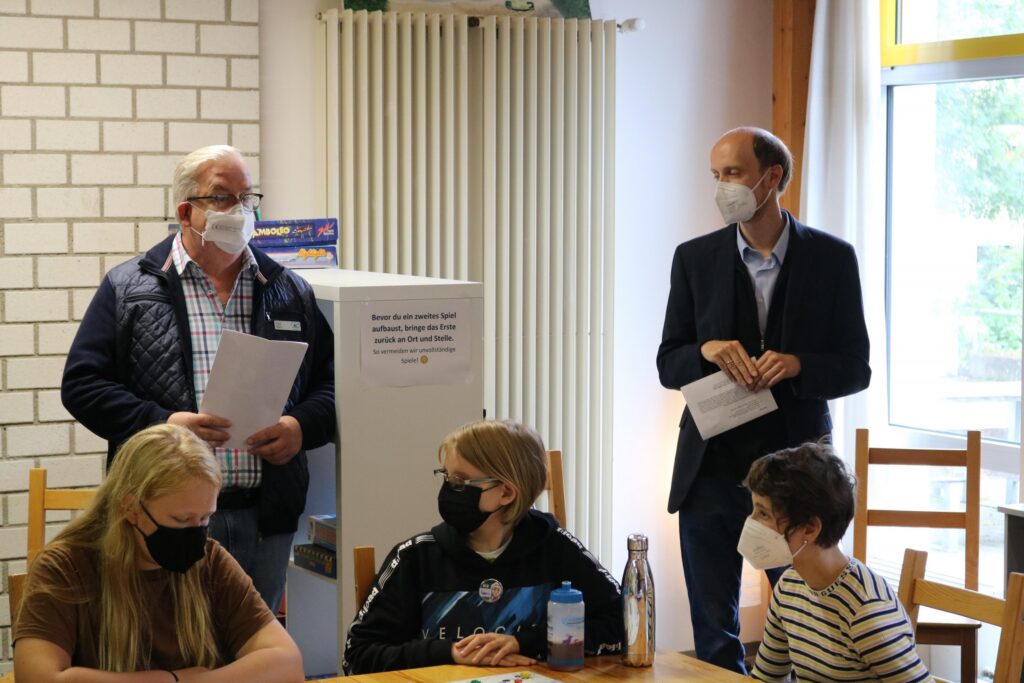Zwei Männer stehen vorne, Schüler sitzen am Tisch, alle tragen Masken.