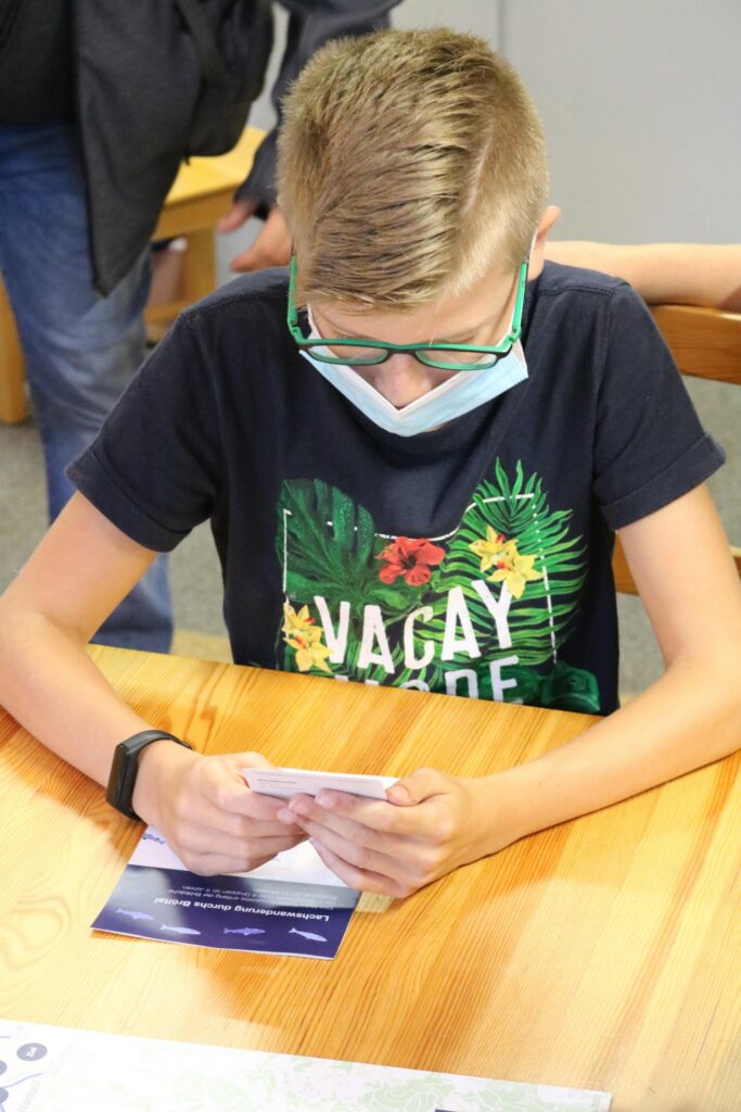Ein Junge mit Brille und Maske sitzt an einem Tisch und schaut auf ein Smartphone.