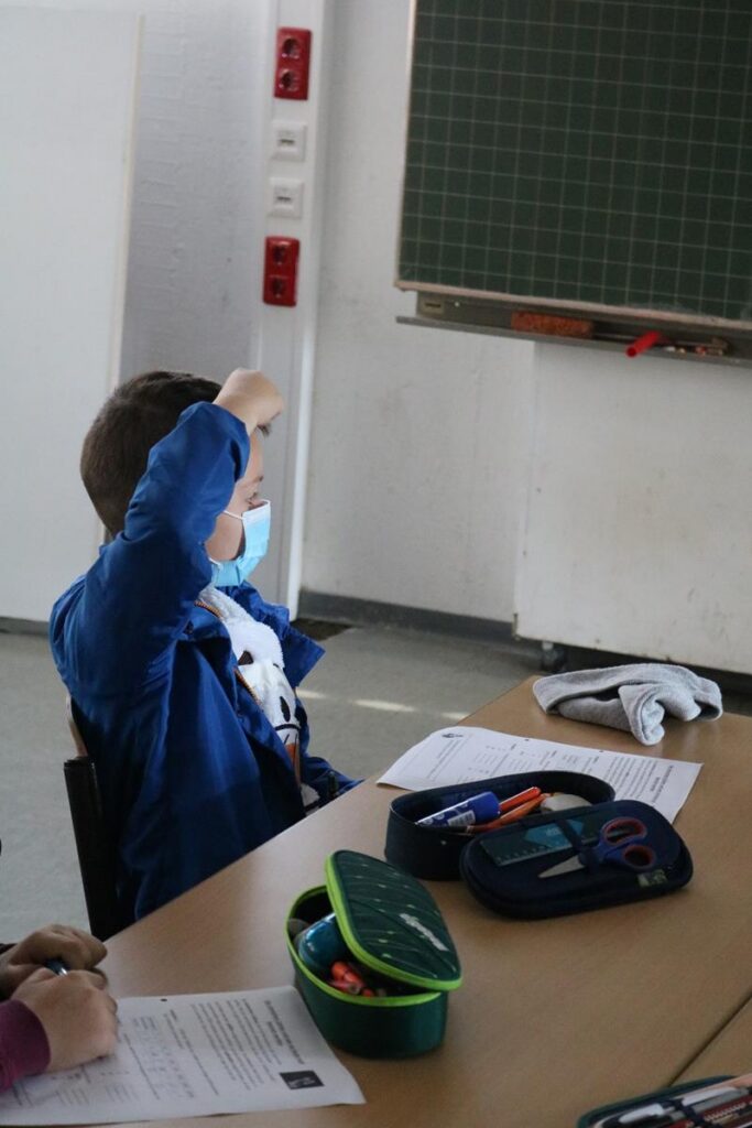 Schüler sitzt an einem Tisch und hebt die Hand. Auf dem Tisch liegen Stifte und Unterlagen.