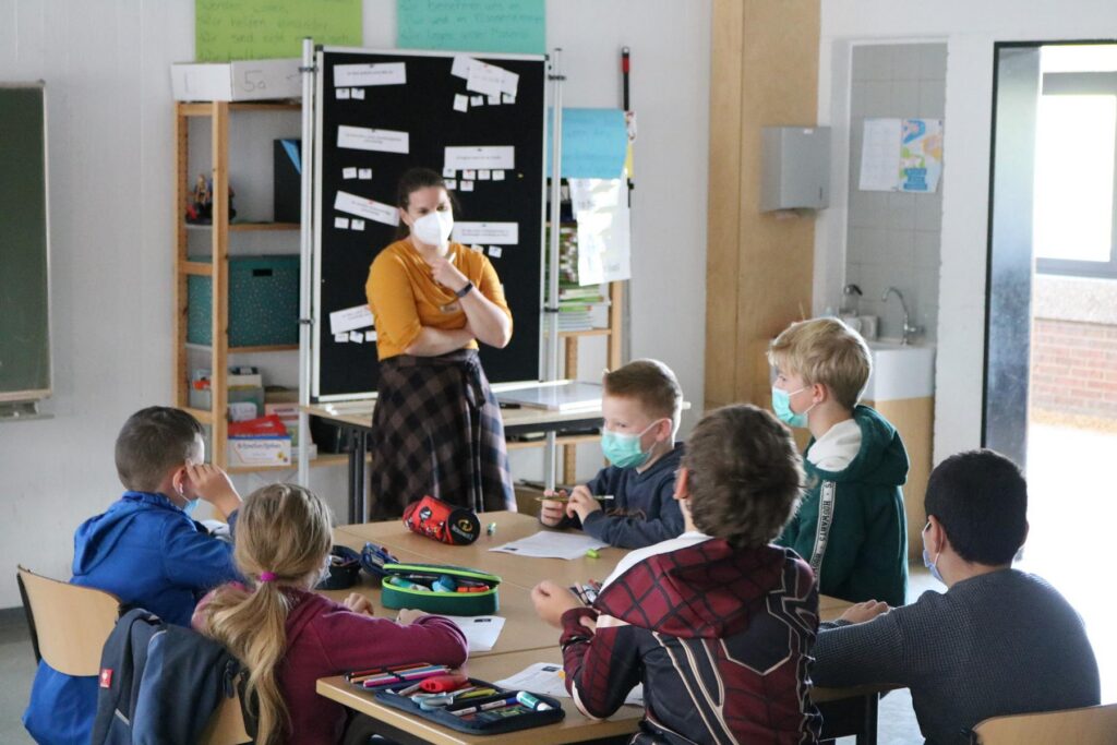 Schüler und Lehrerin im Klassenzimmer bei einer Gruppenarbeit. Alle tragen Masken.
