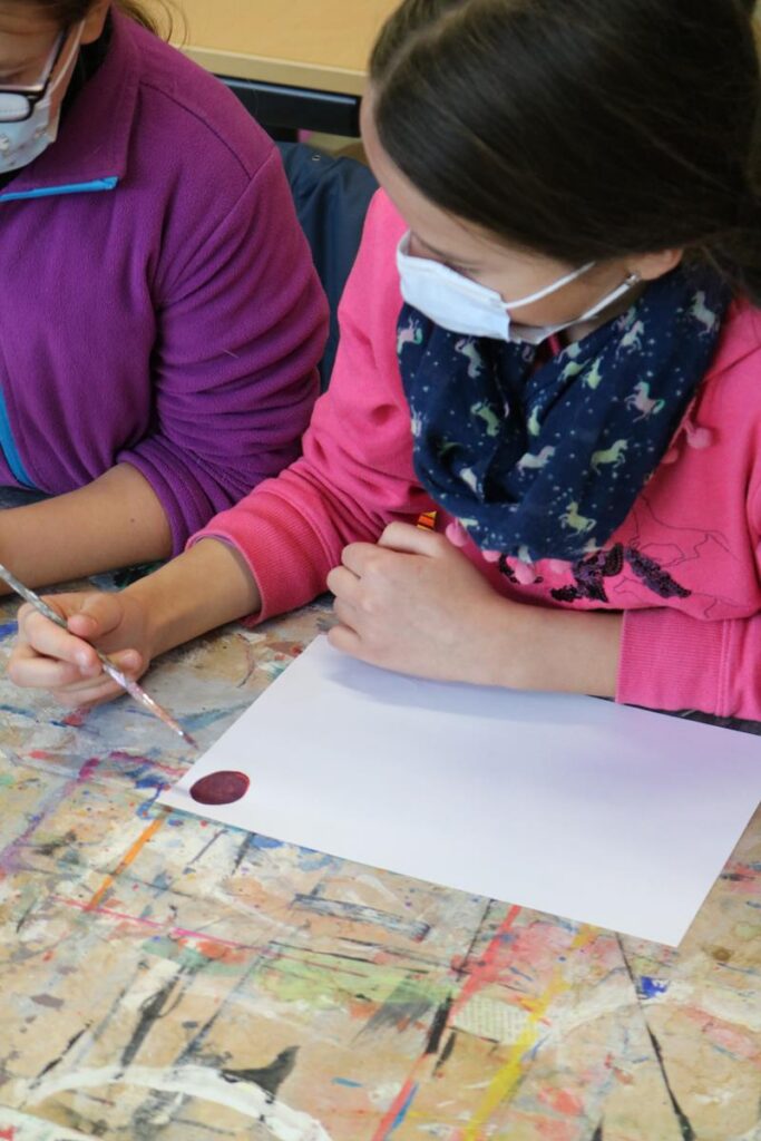 Schülerin malt mit Pinsel auf Papier, Tisch mit Farbresten.