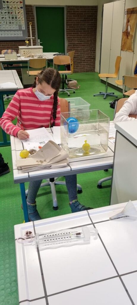 Mädchen mit Maske sitzt am Tisch und arbeitet an einem Experiment.