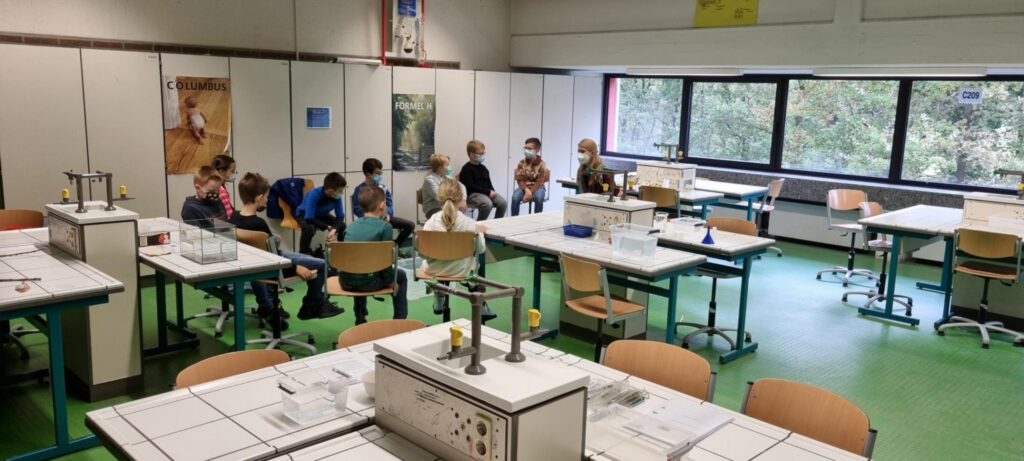 Schüler sitzen in einem Klassenraum mit Tischen und Fenstern.