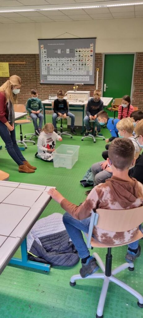 Unterrichtsszene mit Lehrerin und Schülern, die im Klassenraum sitzen und arbeiten.