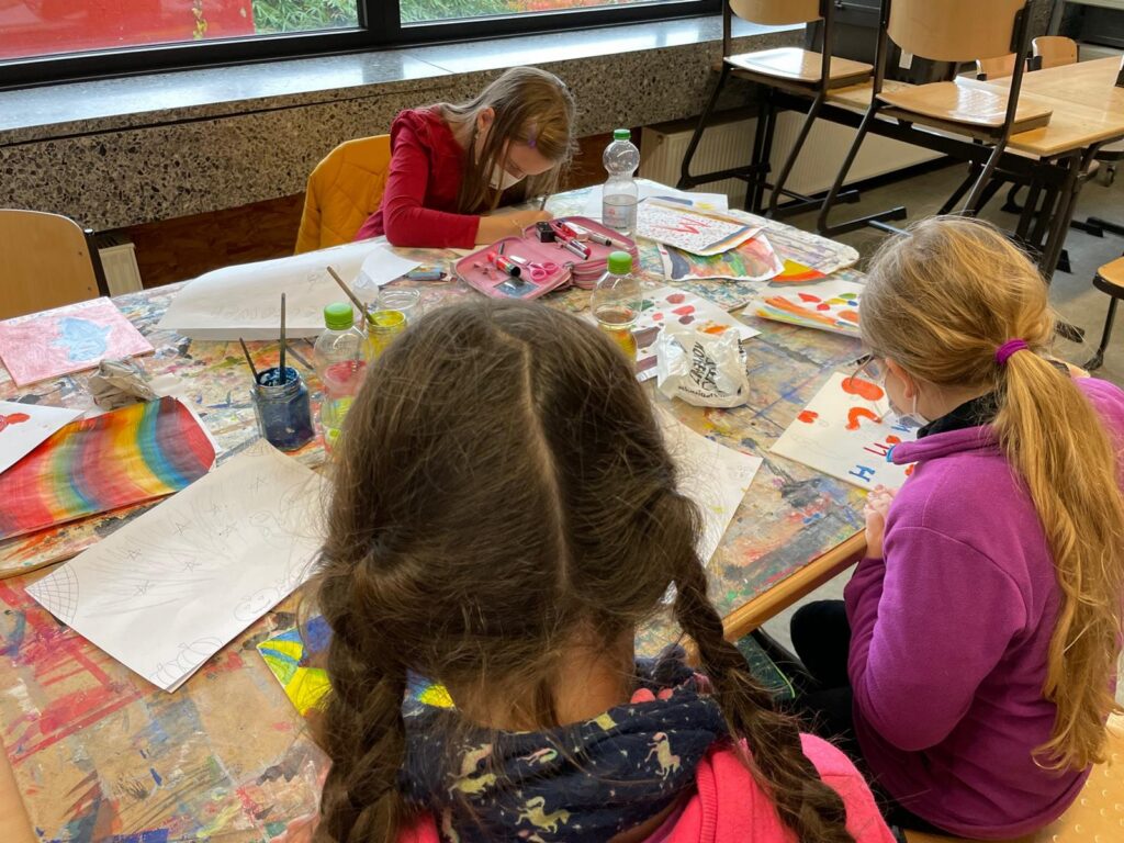 Kinder arbeiten kreativ an einem Tisch mit Malutensilien und buntem Papier.