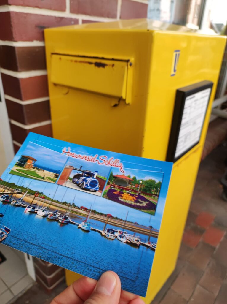Eine Person hält eine Postkarte vor einem gelben Briefkasten.