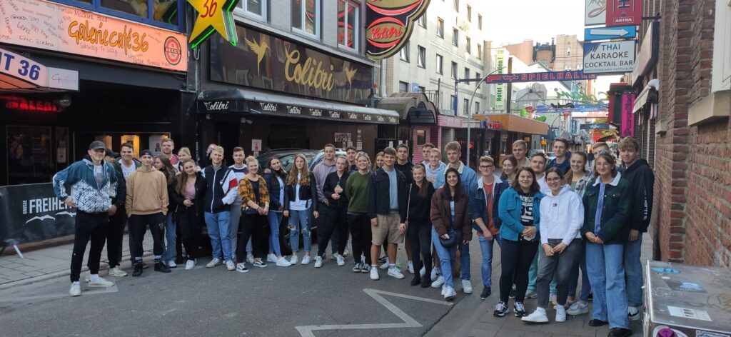 Gruppe von Schülern steht auf einer Straße vor mehreren Geschäften und Bars.