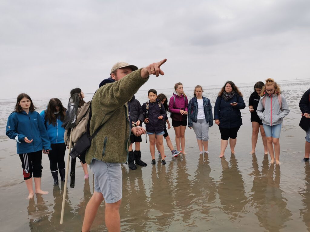 Gruppe von Schülern steht am Strand, ein Lehrer erklärt etwas und zeigt mit dem Finger.