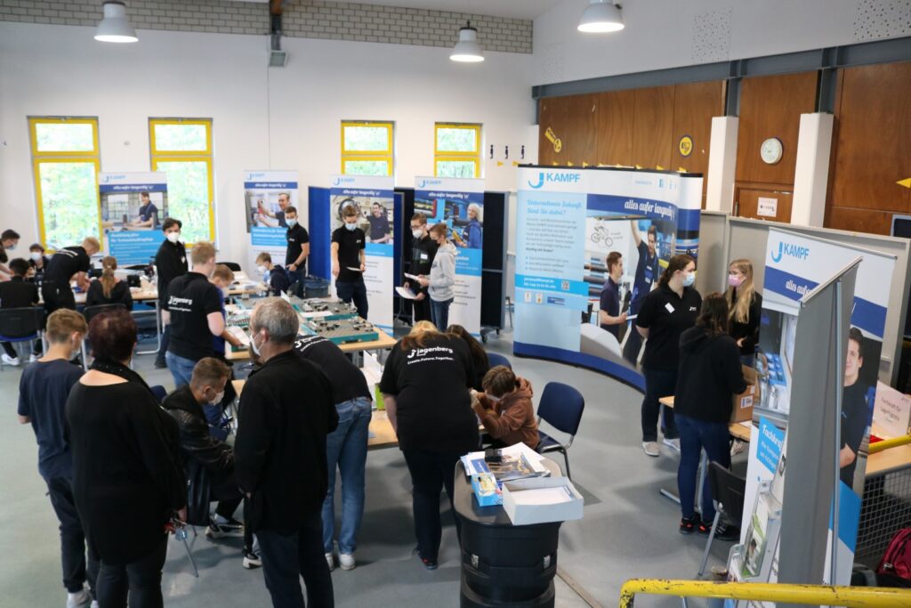 Informationsmesse mit Ständen und Besuchern in einem großen Raum.