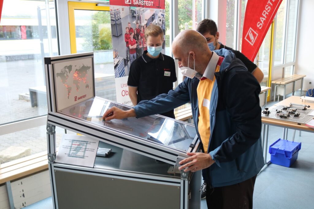 Eine Person untersucht eine Präsentationsanlage, im Hintergrund Mitarbeiter.