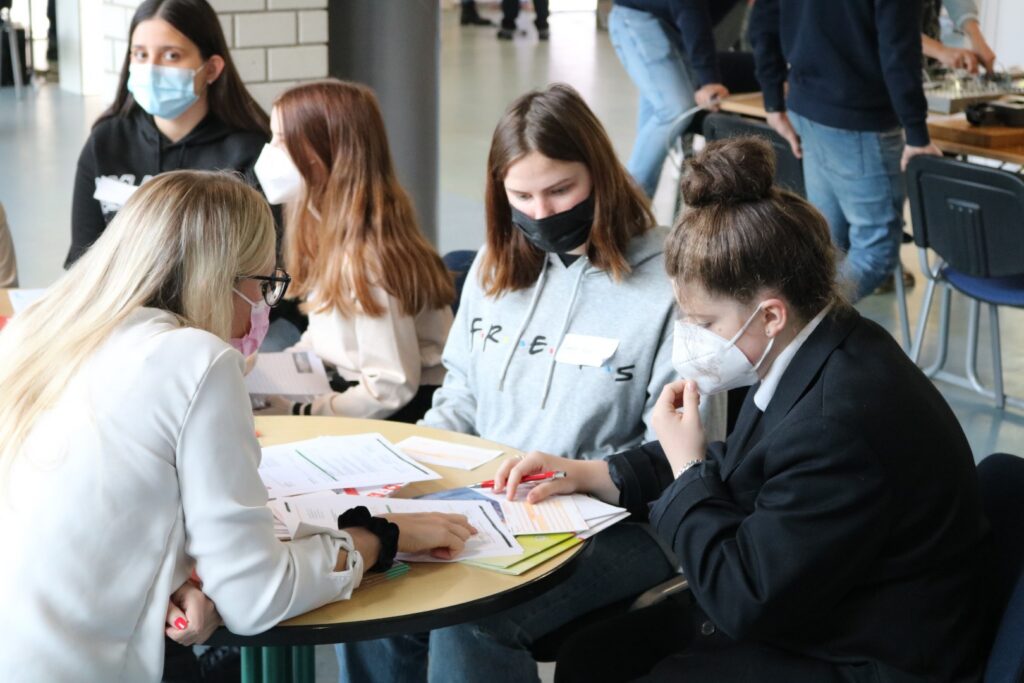 Schülerinnen und Lehrerinnen arbeiten an einem Tisch mit Unterlagen und Masken.