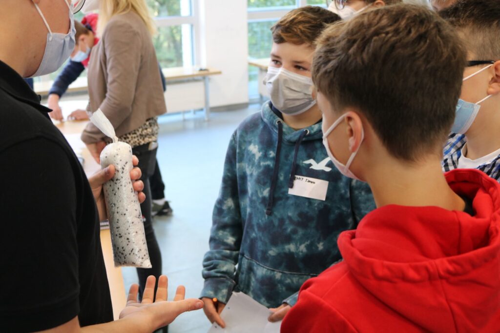 Schüler in einem Klassenraum diskutieren mit Masken und einem Objekt in der Hand.