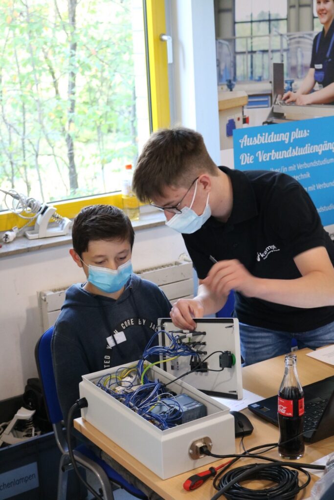 Schüler und Ausbilder arbeiten an einem elektronischen Gerät. Beide tragen Masken.