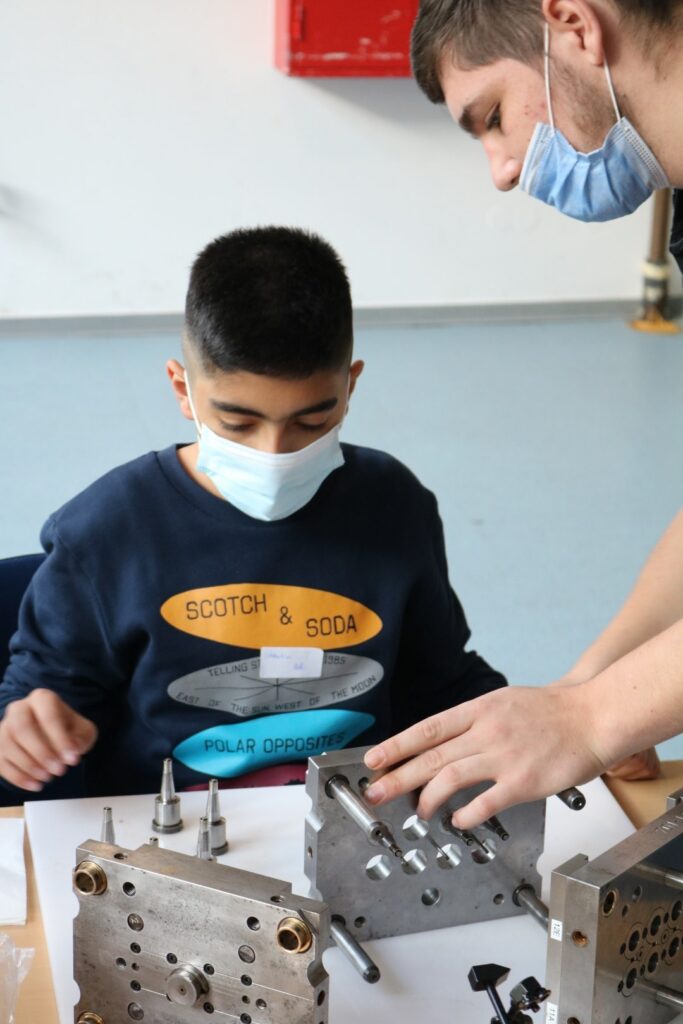 Ein Junge und ein Erwachsener arbeiten an Metallteilen am Tisch. Beide tragen Masken.