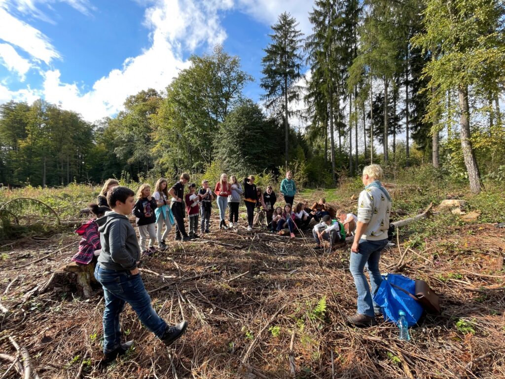 Gruppe von Schülern und Lehrern im Wald, umgeben von Bäumen und Sonnenschein.